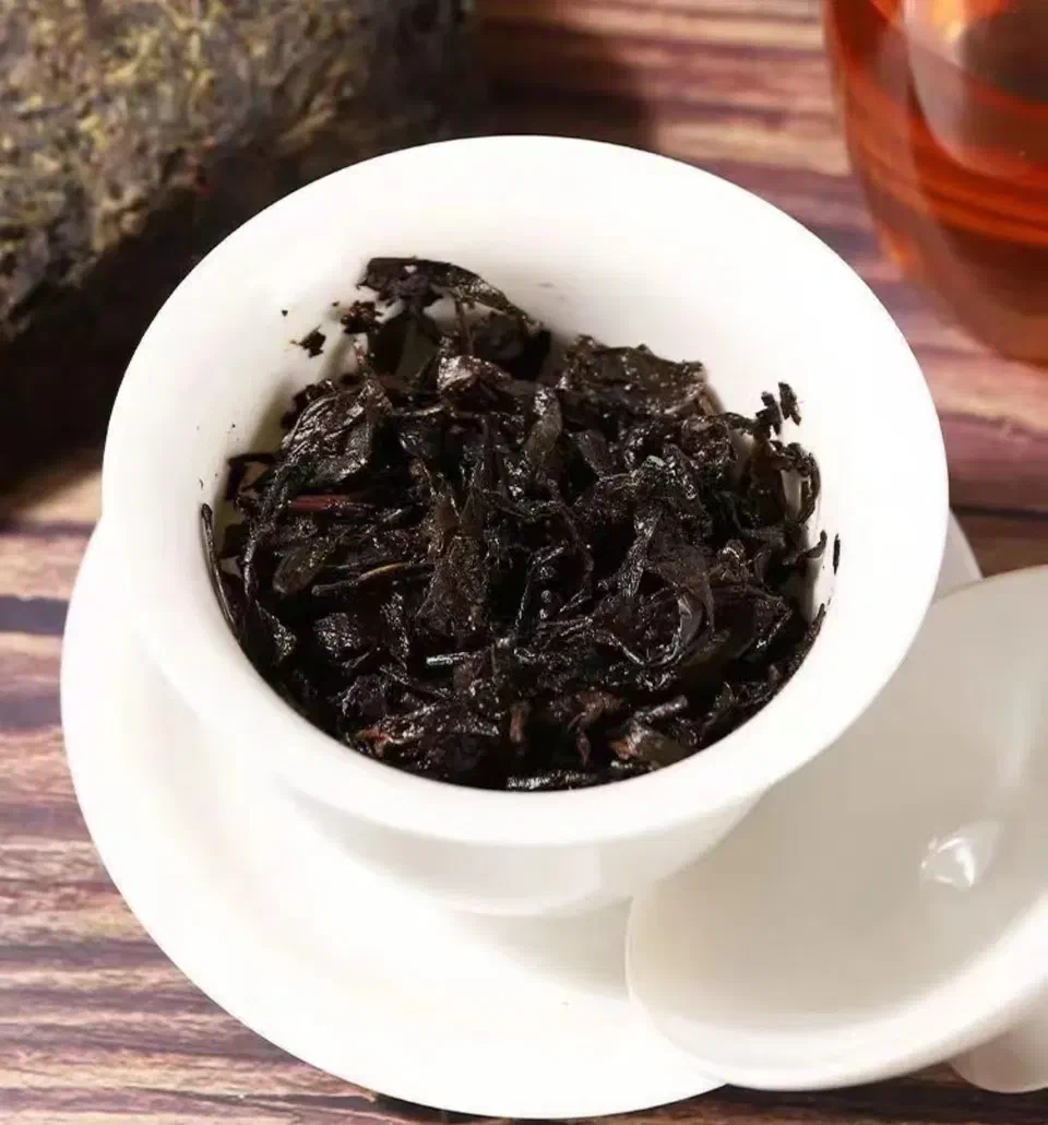 Steeped dark tea leaves in a white gaiwan, with a tea brick and a glass of brewed tea behind it.